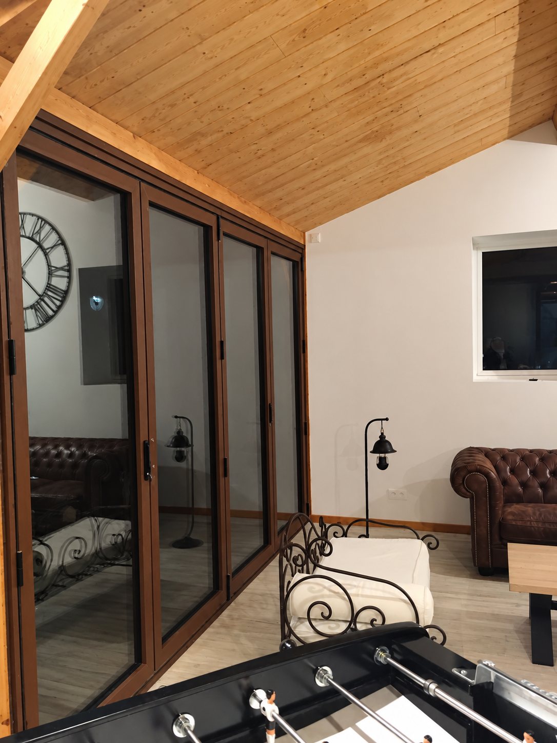 Une grande baie vitrée coulissante à cadre brun donne sur un salon sous plafond en lambris, avec canapé chesterfield, fauteuil en fer forgé et baby-foot au premier plan.