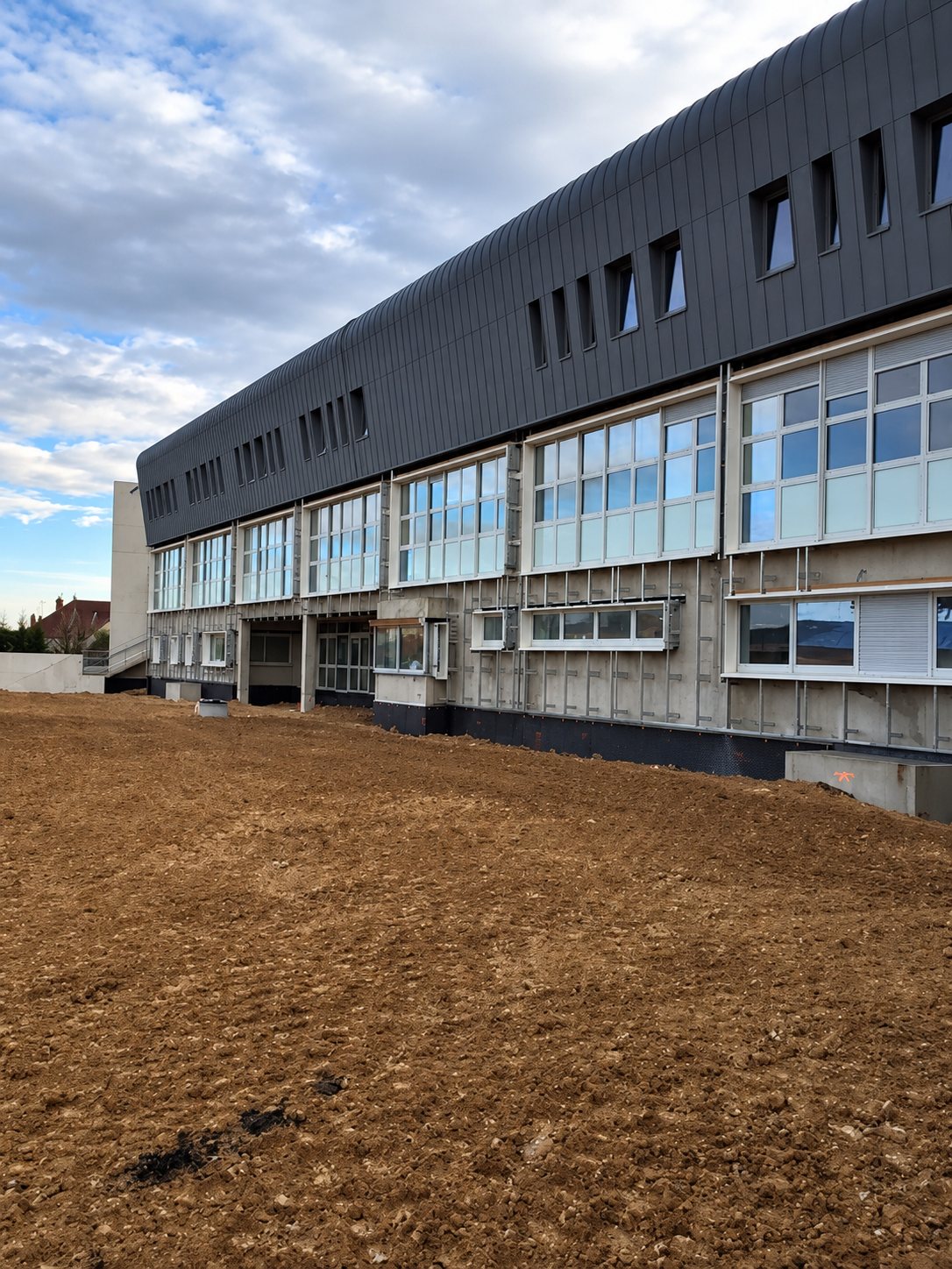 Façade d'un grand bâtiment en rénovation avec bardage métallique sombre à l'étage, longues rangées de fenêtres et terrain de terre nue au premier plan.