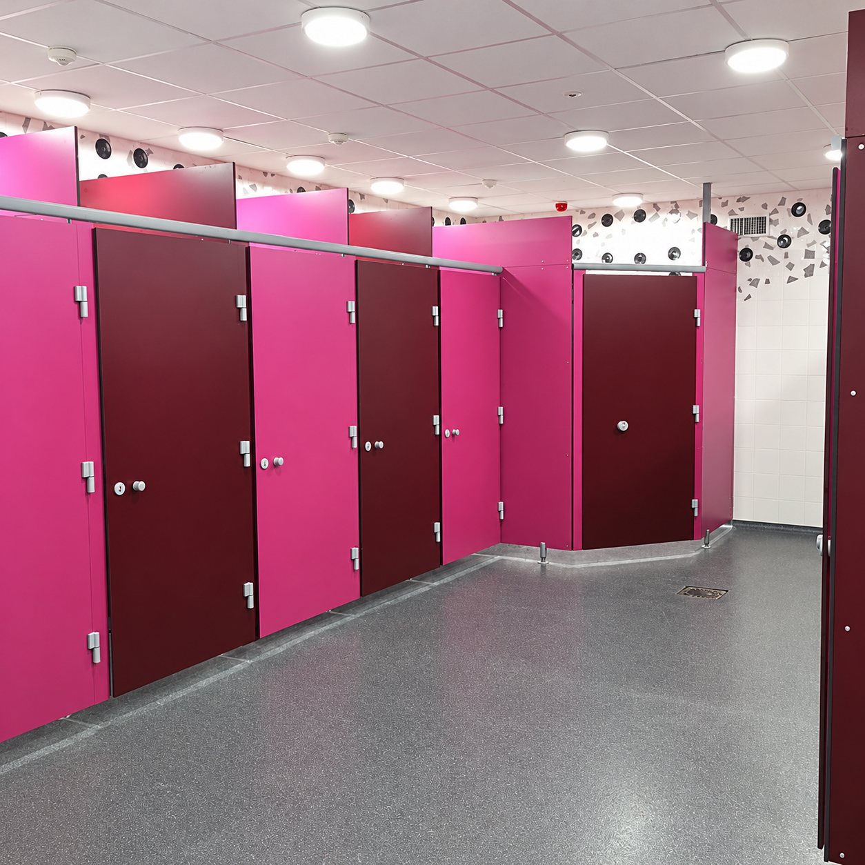 Rangée de cabines de toilettes aux cloisons roses et aux portes bordeaux dans un sanitaire collectif au sol gris moucheté.