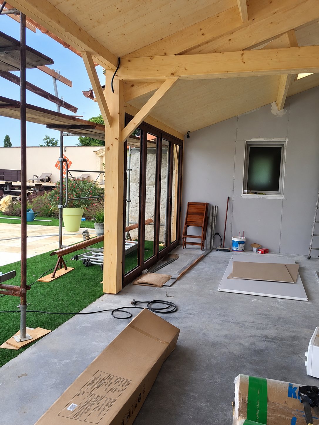 Terrasse couverte en cours de construction avec charpente en bois apparente, grande baie vitrée coulissante, échafaudage et gazon synthétique à côté.