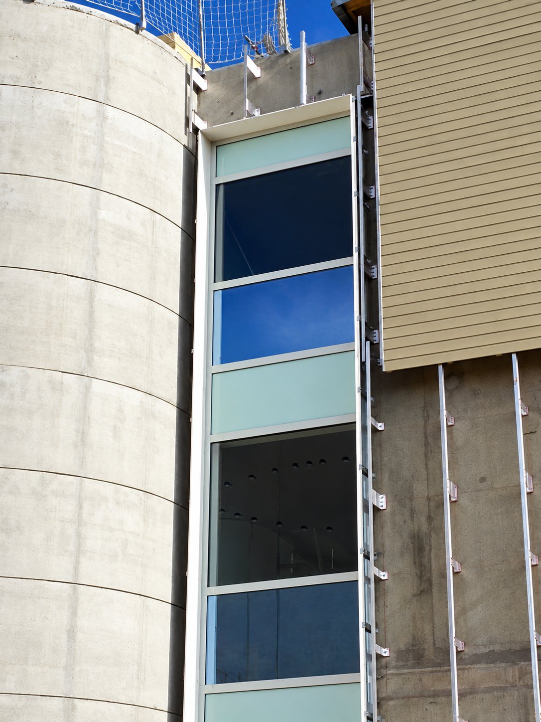 Chantier d'un bâtiment montrant une haute fenêtre vitrée verticale encadrée par un silo cylindrique en béton et un revêtement en bois clair avec des rails de fixation métalliques.