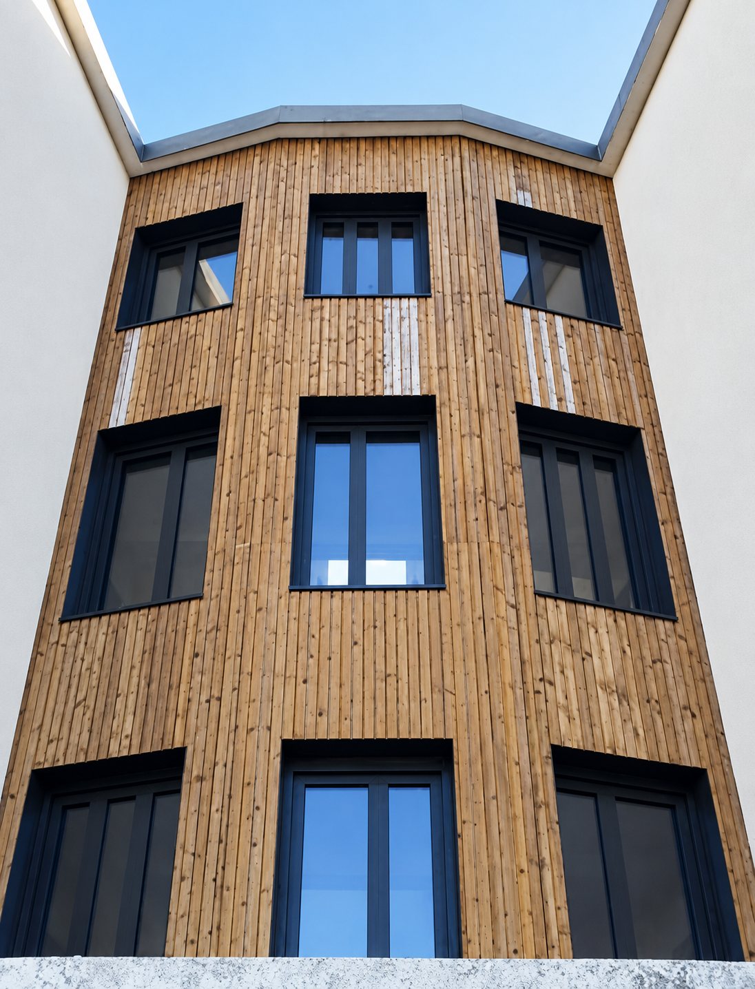 Vue en contre-plongée d'une façade d'immeuble recouverte de bardage bois vertical, percée de neuf fenêtres aux cadres sombres, entre deux murs blancs.