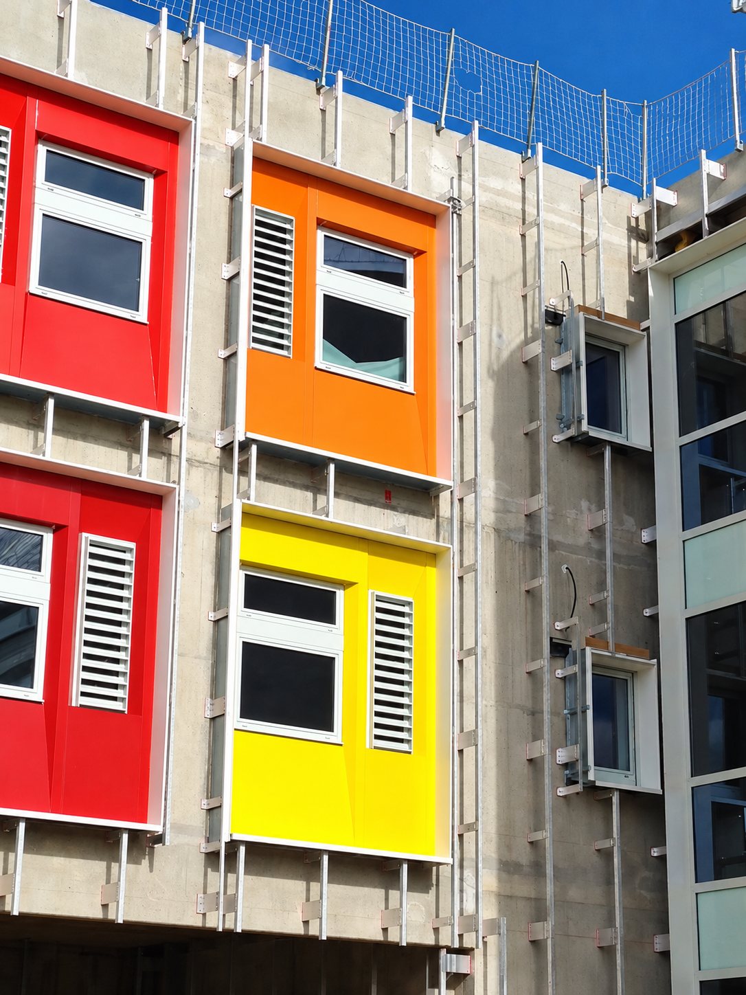 Façade en béton d'un bâtiment en chantier avec des panneaux de fenêtres rouges, orange et jaunes, des échelles fixées au mur et un filet de sécurité en haut.