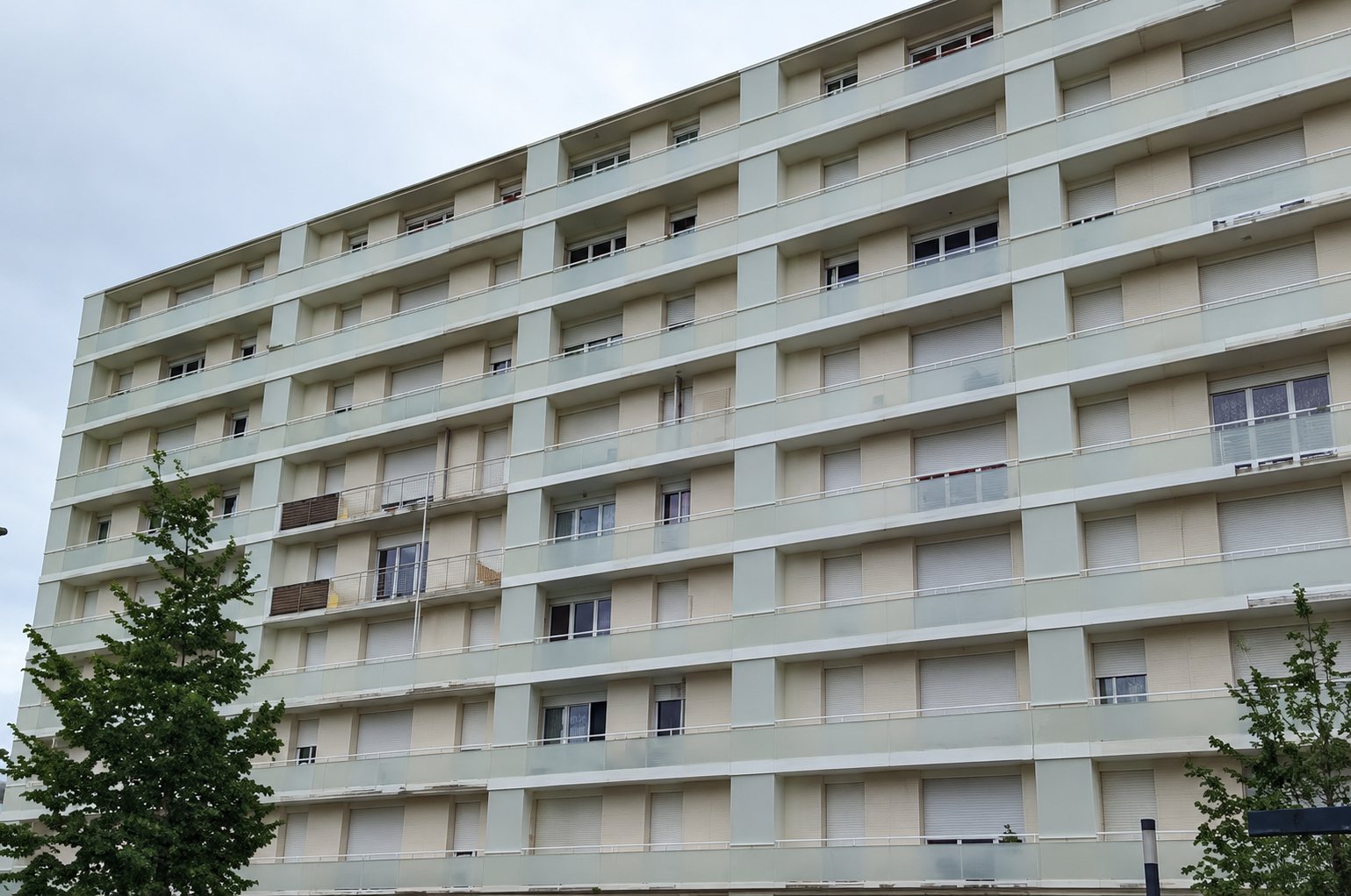 Façade d'un grand immeuble d'habitation clair à nombreux étages, avec balcons et volets roulants, sous un ciel nuageux.