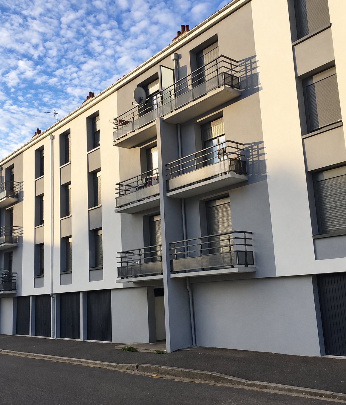 Façade d'un immeuble de trois étages aux murs gris et blancs, équipé de balcons à garde-corps métalliques et de garages au rez-de-chaussée.