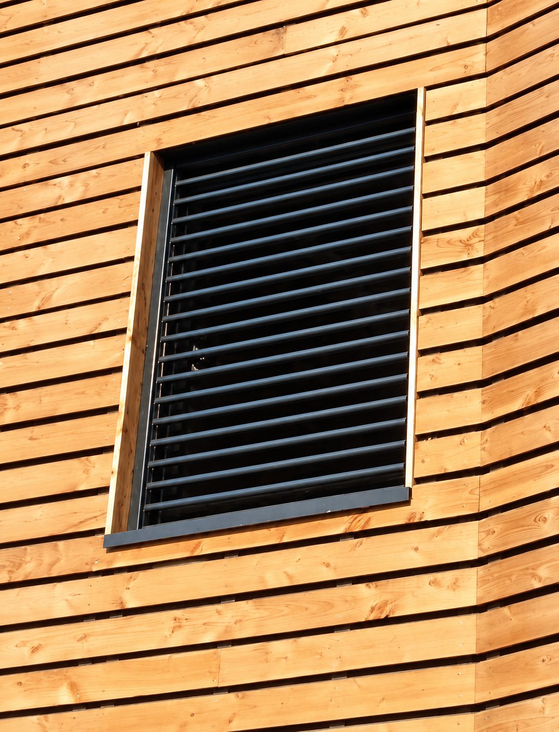 Une fenêtre équipée de lames de persienne grises est encadrée par un bardage en bois clair posé à l'horizontale sur la façade d'un bâtiment.