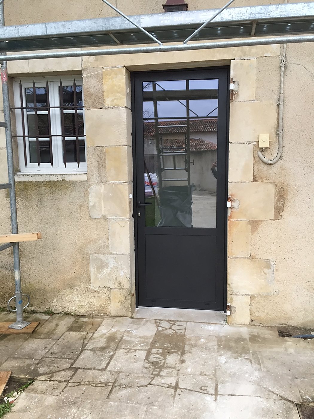 Une porte vitrée en aluminium noir récemment installée dans la façade en pierre d'une maison, à côté d'une fenêtre à barreaux et d'un échafaudage.