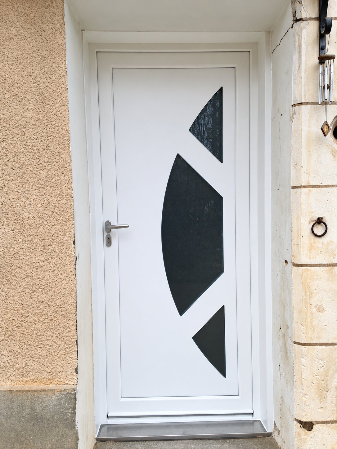 Porte d'entrée blanche à poignée métallique, ornée de trois vitrages sombres en forme de segments, encadrée d'un mur crépi beige et de pierres de taille.