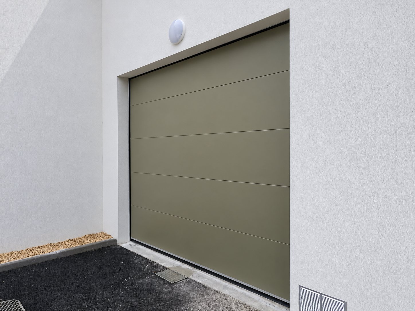 Porte de garage sectionnelle de couleur vert kaki posée dans une façade blanche crépie, avec un luminaire au-dessus et un sol en enrobé devant.