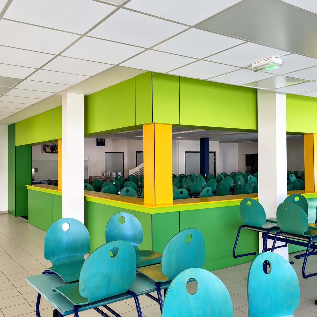 Salle de restauration scolaire aux cloisons vertes et jaunes, avec des chaises turquoise au premier plan et de nombreuses tables en arrière-plan.