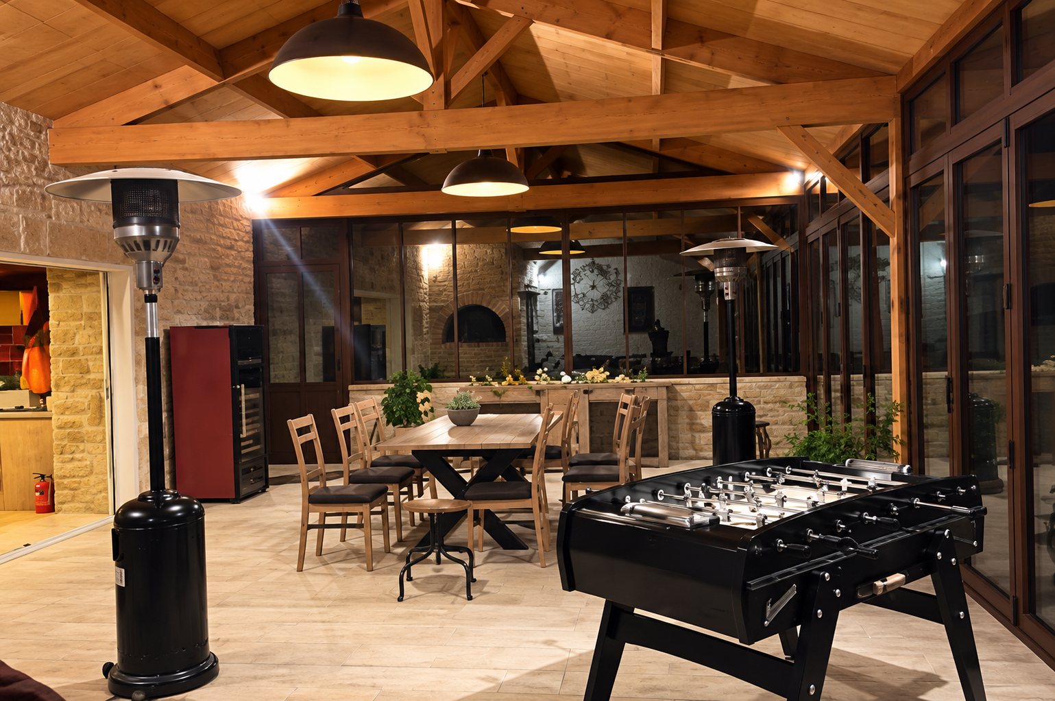 Grande salle aux poutres apparentes équipée d'un baby-foot, de tables et chaises en bois et de lampes suspendues, éclairée en soirée.