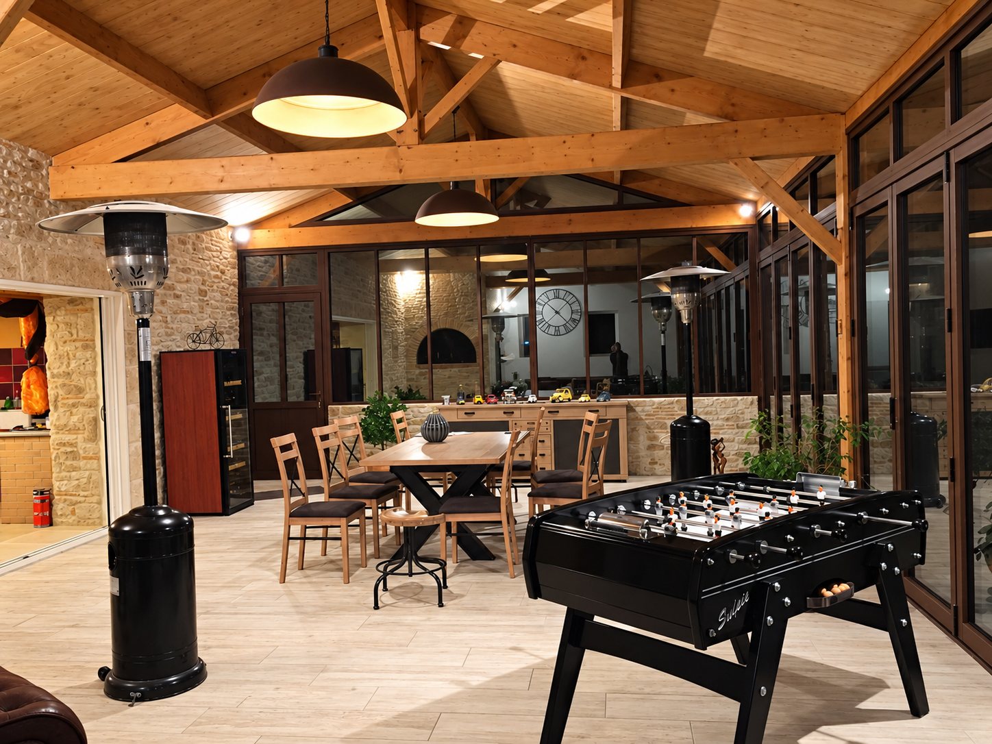 Une grande pièce aux murs en pierre et à la charpente apparente en bois, meublée d'une table avec chaises, d'un baby-foot noir et de chauffages d'extérieur, éclairée le soir.