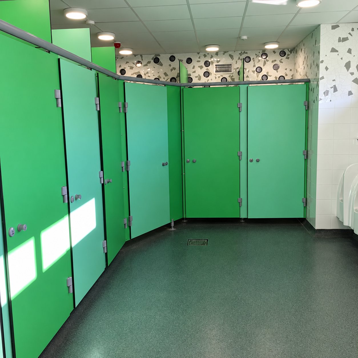 Intérieur de sanitaires récemment aménagés avec des cabines aux portes vertes, des urinoirs à droite et un mur décoré de motifs graphiques.